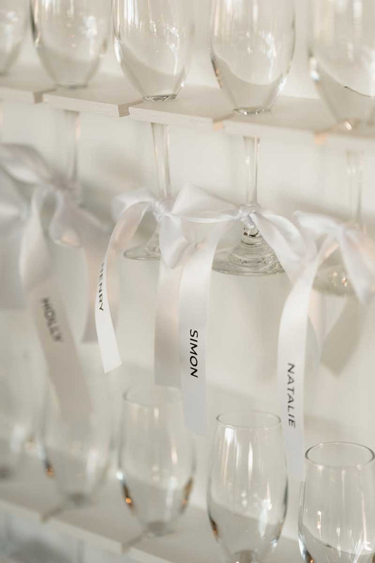 A close-up of several clear wine glasses hanging from white shelves, each tied with a white ribbon with names on them, wedding escort card