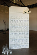 A white vertical wedding reception wall display holding many clear wine glasses tied with white ribbons, decorated with white flowers at the top and candles on the floor, photographed at the entrance to wedding reception