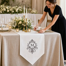 A woman in a black dress adjusts a white table runner with a black ornate monogram 'M' on a beautifully set wedding table with white flowers, gold candlesticks, and wine glasses.