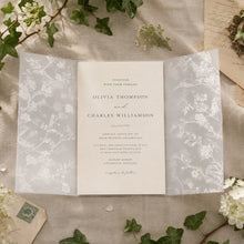 A vintage-style wedding invitation card displayed in a semi-transparent floral lace envelope, styled on a textured linen fabric with green ivy leaves and old paper props.