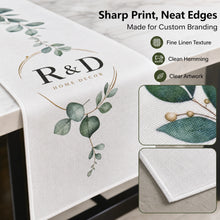 A white table runner with a green eucalyptus leaf print and a custom 'R&D Home Decor' logo, draped over a table. The image includes marketing text on the right highlighting its sharp print, neat edges, fine linen texture, clean hemming, and clear artwork, along with two inset close-up photos of the fabric details.