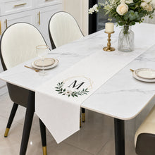 A white table runner with a monogram 'M' and green leaf wreath design, styled on a white marble dining table with chairs, plates, and a vase of flowers.