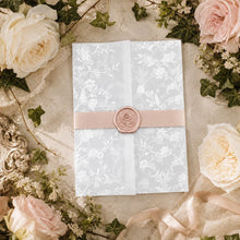 A square white envelope or card with a delicate floral damask pattern, tied with a pale pink ribbon and sealed with a round pink wax seal, styled on a beige fabric background with roses, greenery, and lace.