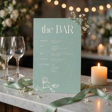 A vertical bar menu card in a muted sage green color stands on a marble table runner with green ribbon, small white flowers, wine glasses, and a lit candle. The card has elegant white typography reading 'the BAR' with cocktail and wine list details below from the Aleisha Collection.