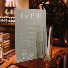 A printed bar menu card titled "the BAR" with white magnolia flower design, stands on a wooden bar counter in a rustic bar setting, with a clear glass and bar fixtures blurred in the background, Aleisha Collection