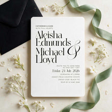 The Aleisha Collection, a classic wedding invitation card with rounded corners, featuring black serif text. The card is styled in a flat lay with a black envelope, small white flowers, and a pale green ribbon on a light beige surface
