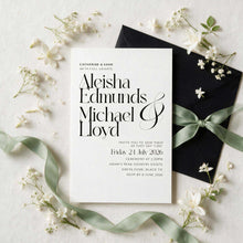 A styled flat-lay of a wedding invitation card with elegant black typography, placed on top of a black envelope tied with a pale green ribbon, surrounded by small white flowers on a light textured background.