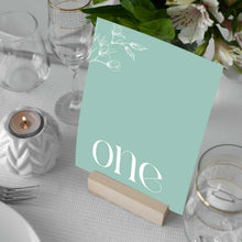 A sage green wedding table number card standing on a small wooden block at a decorated table setting with white magnolia flowers, clear glassware, and a white tablecloth, Aleisha Collection.