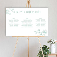A vertical wedding seating chart print with the title 'OUR FAVOURITE PEOPLE' in mint green, displayed on a wooden easel. The white print features delicate magnolia floral illustrations in the corners and lists names by generations.