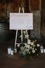 A wedding reception seating chart sign reading 'OUR FAVORITE PLACE' displayed on a white easel, surrounded by white magnolia flowers and candles in a rustic venue setting.