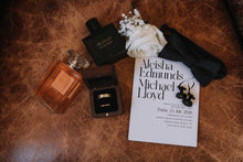 A styled flat-lay of wedding and gift items on a brown leather surface, including a black Michael Kors perfume bottle, a ring box with a gold band, a white rose, a black bow tie, and a wedding invitation card