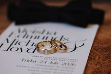 Two gold wedding bands resting on a white wedding invitation card, with a blurred black bow tie in the background on a brown textured surface.
