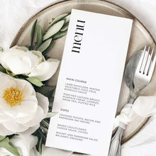 A vertical white wedding menu card with elegant black typography, styled on a round ceramic plate with white magnolia flowers, green leaves, and silver flatware.