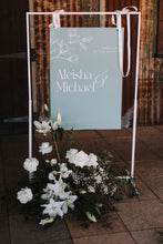 A wedding reception sign reading 'Aleisha & Michael' in white script, a handdrawn magnolia flower, on a sage green background, displayed in a white metal frame with white flowers and greenery at the base on a tiled floor.