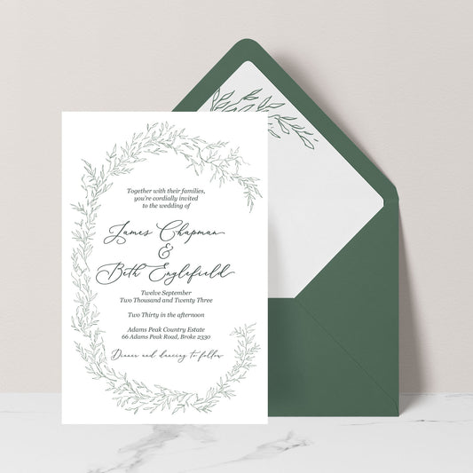 A wedding invitation card with a delicate green botanical wreath border and elegant script text, displayed in front of a dark green envelope on a light neutral background.