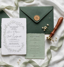 A styled flat lay of a wedding invitation suite on white satin fabric, featuring a white invitation card with a gray floral wreath border, a sage green RSVP card, a dark green envelope with a gold wax seal, and a wooden-handled seal stamp.