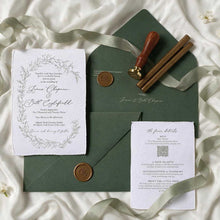 A styled flat-lay of a wedding invitation suite on a white satin background. The set includes a white paper invitation card with a delicate green floral wreath border and elegant script, a dark forest green envelope, a wooden-handled wax seal stamp, two wooden wax seals with gold-toned caps, and a white RSVP card.