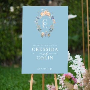bridgerton inspired vintage floral wreath with monogram welcome sign blue and peach florals sitting on easel in garden 