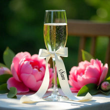 Champagne flute with a white ribbon and 'Sam' inscription, surrounded by pink flowers outdoors.