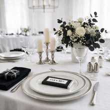 A styled wedding table setting with white plates, silver-rimmed chargers, a black napkin with a wrapped gift, silver salt and pepper shakers, a floral centerpiece with white roses and black eucalyptus, and candlesticks on a white tablecloth.