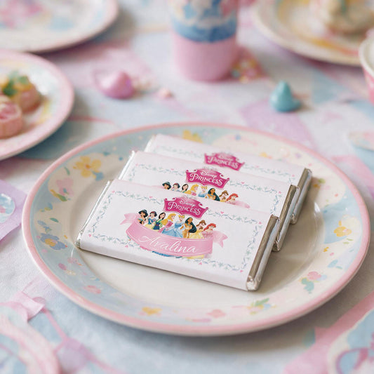 Three wrapped chocolate bars with Disney Princess labels are stacked on a pastel floral plate at a tea-time table setting.