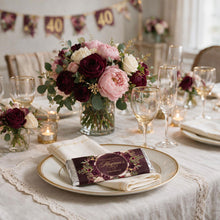 A styled wedding table setting with a floral centerpiece in burgundy, pink, and white roses, gold-rimmed plates, clear wine glasses, and a personalized chocolate bar on a cream tablecloth.