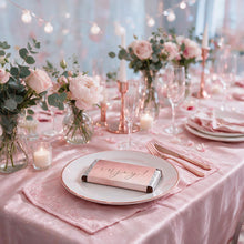 A styled pink table setting with a satin pink tablecloth, white plates, blush pink napkins, rose gold cutlery, and floral centerpieces with pink roses and eucalyptus. A wrapped chocolate bar with the word 'enjoy!' in script sits on the front plate.