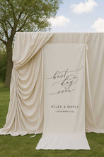 fabric wedding welcome sign featuring words best day ever and couple name and date in front of draping backdrop at garden ceremony