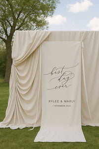 fabric wedding welcome sign featuring words best day ever and couple name and date in front of draping backdrop at garden ceremony