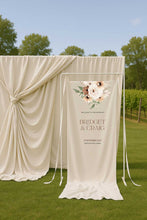 Wedding welcome sign with bohemian floral design and text in an outdoor setting  draped fabric backdrop outdoors.