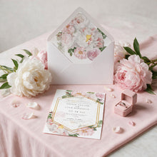 A styled wedding invitation suite on a blush pink fabric surface, featuring a white envelope with a floral liner, a matching printed invitation card, scattered pink flower petals, large white and pink peonies, and a small pink ring box with a ring.