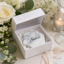 Marble tile coaster with name decal, inside a white box, surrounded by flowers and a candle at a wedding reception, wedding favor