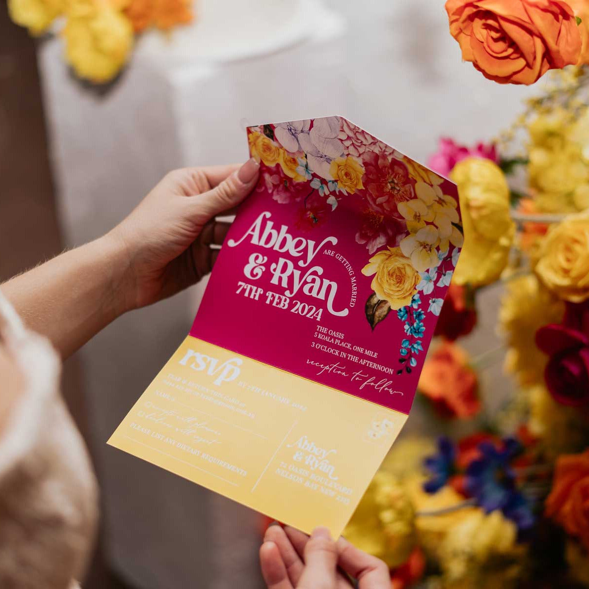 bride holding a colourful floral all-in-one wedding invitation with floral design against a vibrant flower wall.
