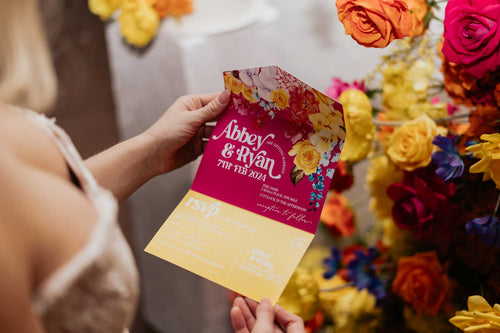 bride holding a colourful floral all-in-one wedding invitation with floral design against a vibrant flower wall.