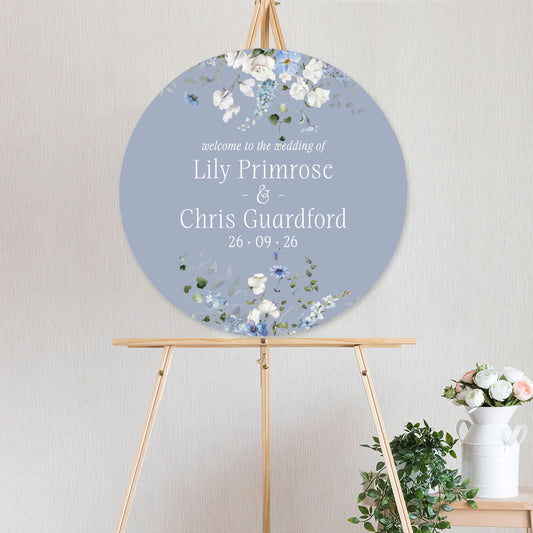 Round wedding sign with white and blue floral design primrose spring collection and text on a stand against a light background