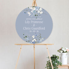 Round wedding sign with white and blue floral design primrose spring collection and text on a stand against a light background