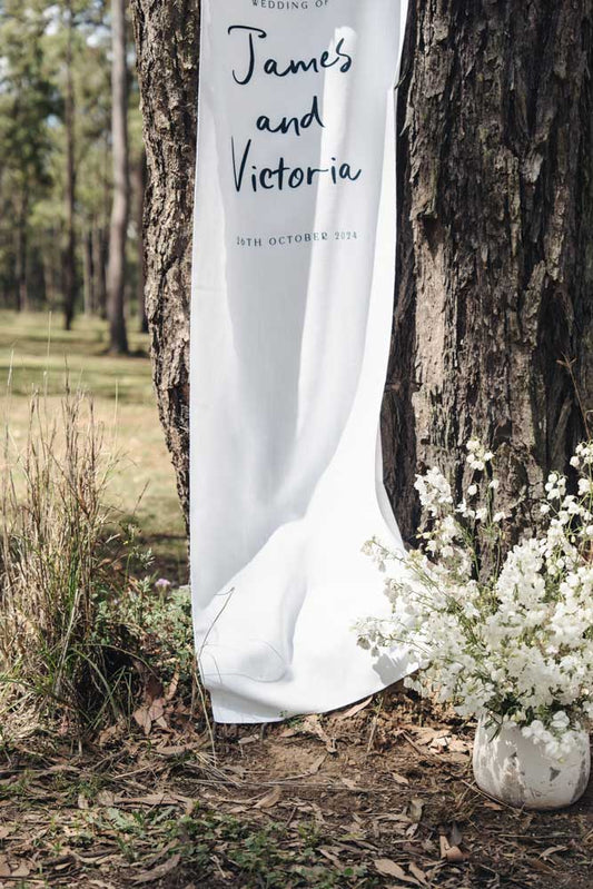 White wedding curtain with 'James and Victoria' text draped over a tree in a forest setting.
