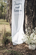 White wedding curtain with 'James and Victoria' text draped over a tree in a forest setting.