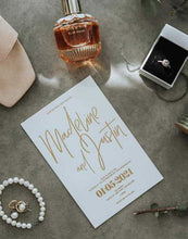Modern wedding invitation card with gold text on a concrete surface with perfume, ring, and jewelry.