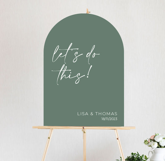 An arch shaped green welcome sign with white text that reads 'Let's do this!' on foamboard or acrylic, mounted on a wooden easel.