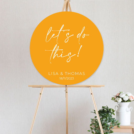 An circle shaped yellow welcome sign with white text that reads 'Let's do this!' on foamboard or acrylic, mounted on a wooden easel.