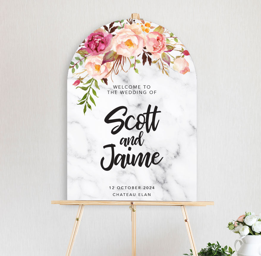An arch shape welcome sign featuring a floral arrangement with peonies in pink and peach hues with marble background with a script font for engagement, wedding, birthday