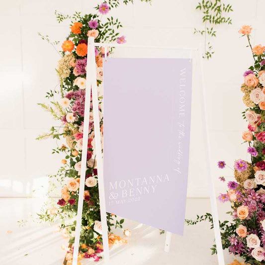 An elegant modern welcome sign in purple and white, mounted on a white stand, with decorative flowers in the background