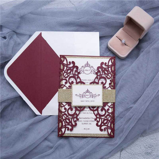 Gate-fold laser-cut wedding invitation featuring a swirl design in burgundy with a gold glitter belly band, backing card and envelope liner