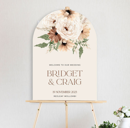 a bohemian floral welcome sign featuring peonies and daisy in autumn brown and neutral tones, diecut to arch shape and sitting on timber easel
