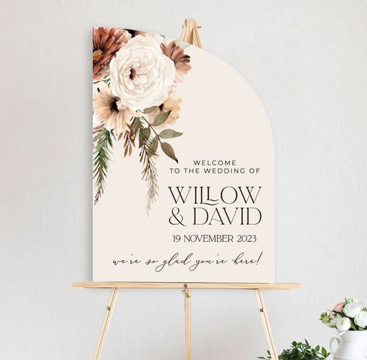 a bohemian floral welcome sign featuring peonies and daisy in autumn brown and neutral tones, diecut to half arch (sail) shape and sitting on timber easel