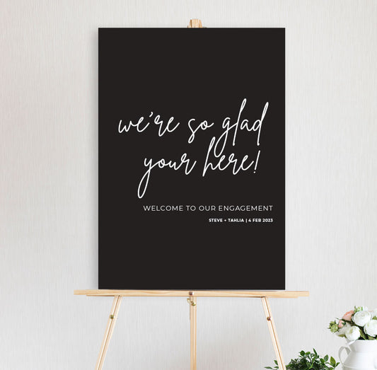 black and white glad you're here fun modern wedding welcome sign sitting on a timber easel engagement party, birthday