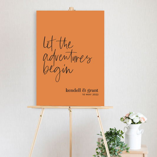 let the adventures begin black and terracotta colour modern script welcome sign on timber easel